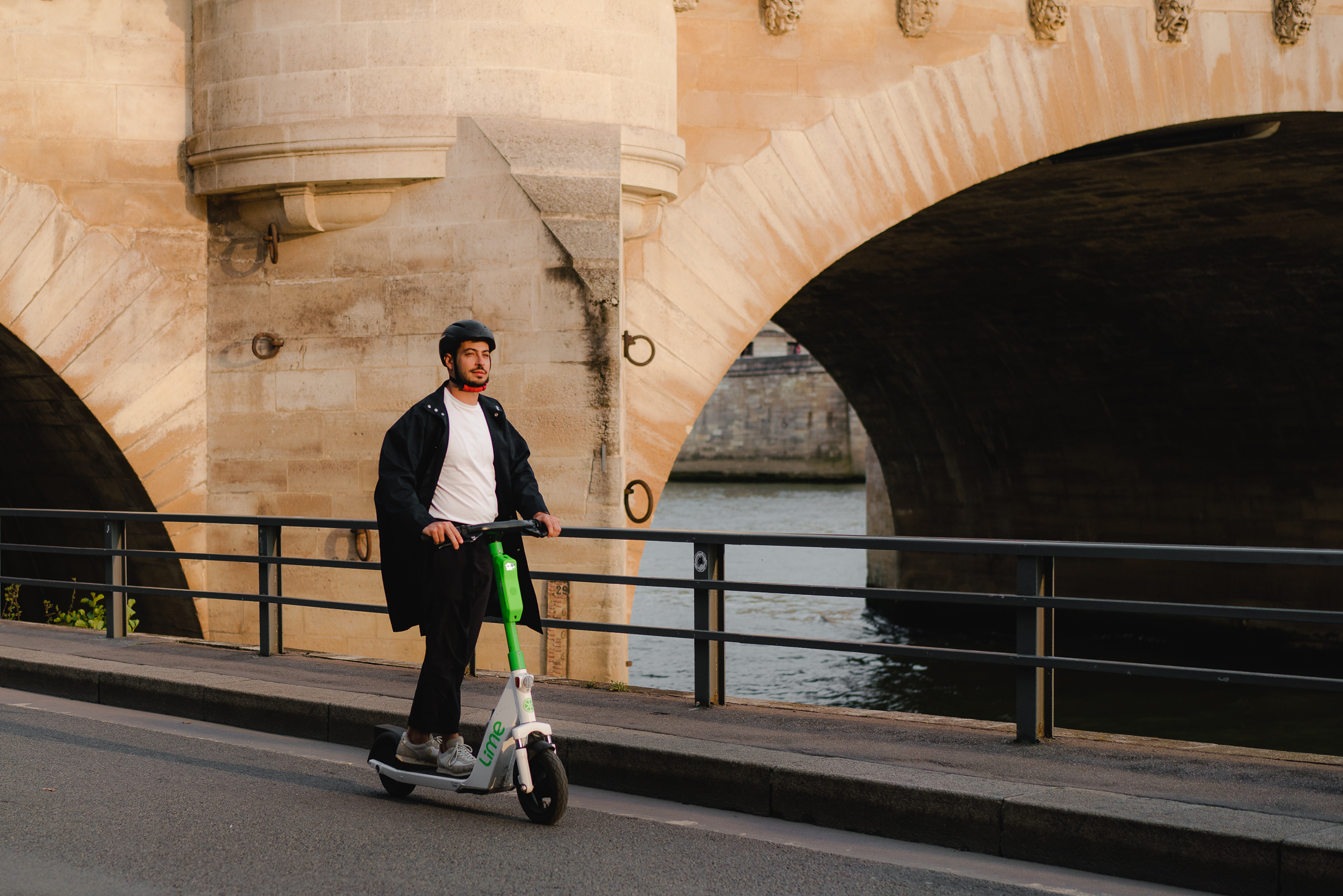 Man riding a Lime e-scooter on a road in front of a bridge crossing water, photo courtesy of Lime