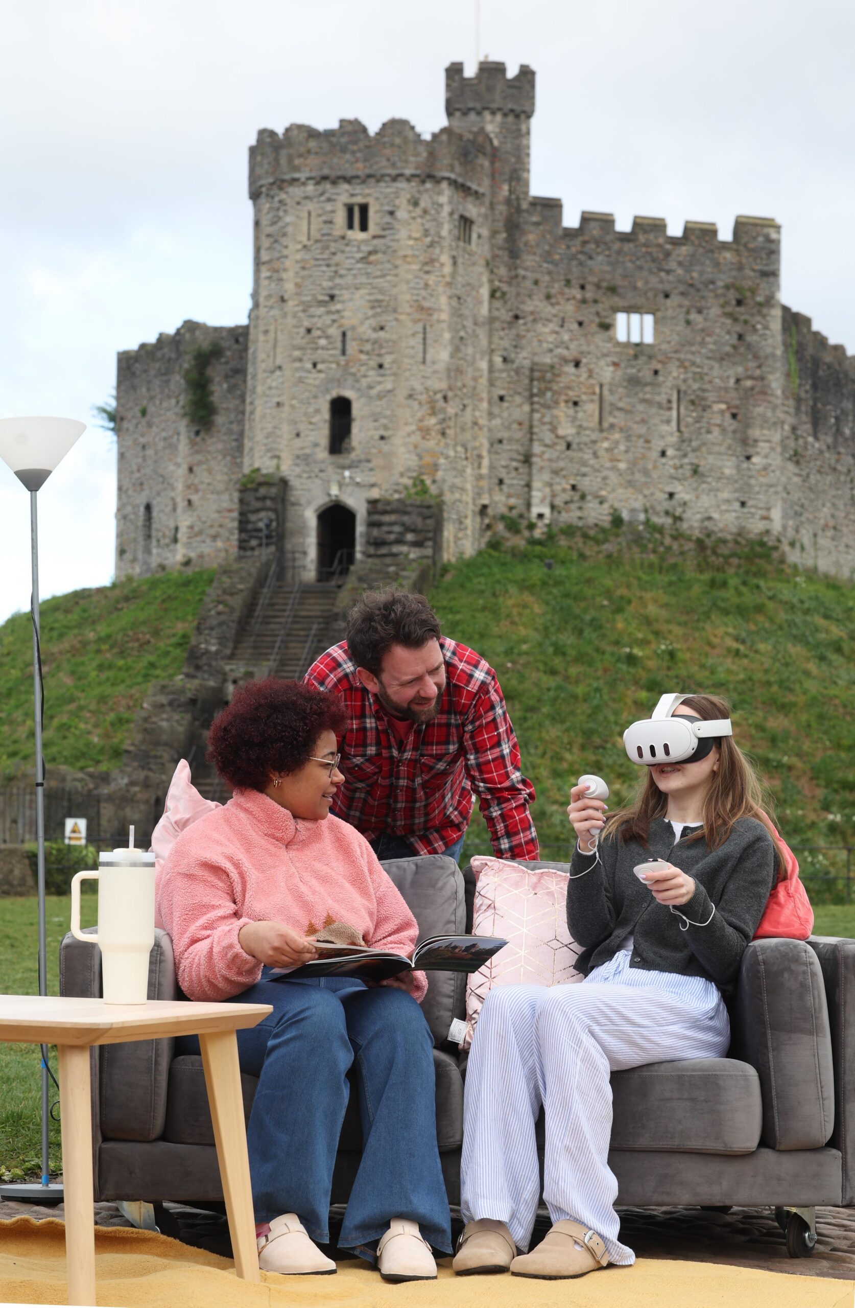 In front of a Welsh castle, two adults sit supervising a child in a VR headset
